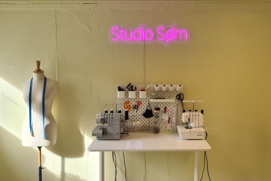 Gul vegg med Studio Søm i neonlys og sysaker på et bord